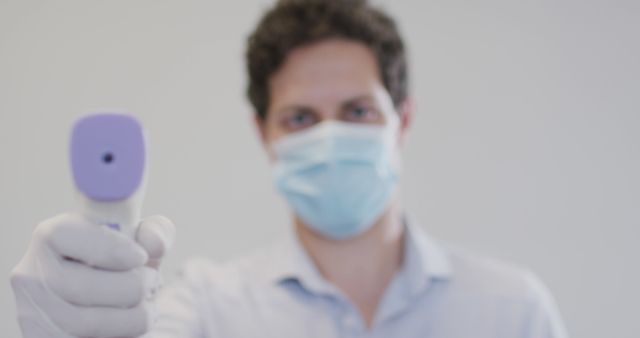 Healthcare professional is holding an infrared thermometer, performing a temperature check. He is wearing a mask and gloves, suggesting a focus on safety and health precautions. This image is ideal for use in articles and marketing materials regarding pandemic safety measures, healthcare protocols, and COVID-19 awareness campaigns.