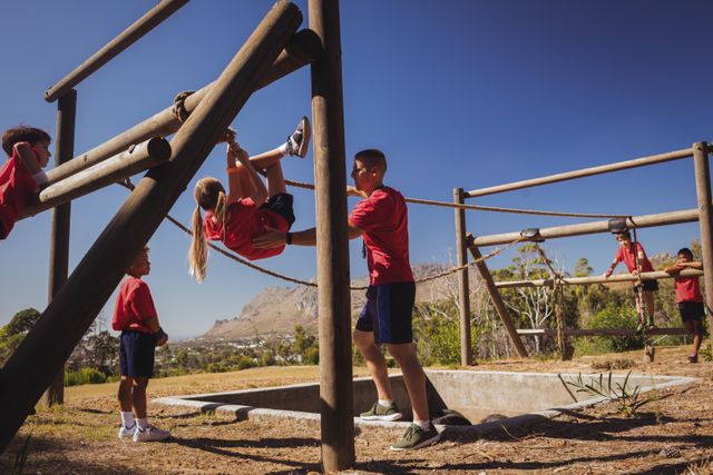 Trainer assisting a girl in navigating an obstacle course at a boot camp. Other children are participating in the background. Ideal for use in articles or advertisements about children's fitness, teamwork, outdoor activities, and summer camps.