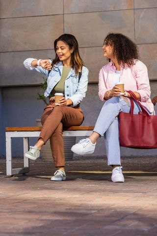 Two african american friends sitting on bench holding coffee and checking wristwatch