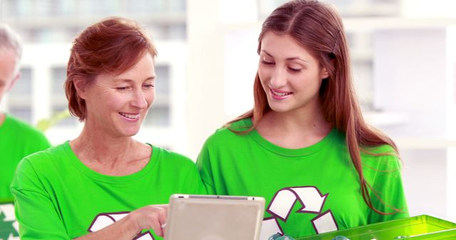 Two female volunteers are smiling while discussing recycling initiatives. Both wearing bright green shirts with a recycling logo, indicating their involvevement in environmental activities. They are likely planning recycling strategies or educating others about sustainability using a tablet. Perfect for promoting recycling programs, environmental awareness, and community volunteer events.