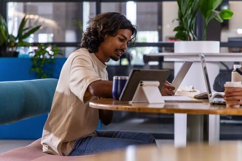 Indian man working on tablet and laptop in modern coworking lounge with coffee and plants