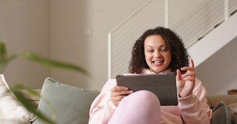 Smiling african american woman in pink loungewear using tablet on cozy living room couch