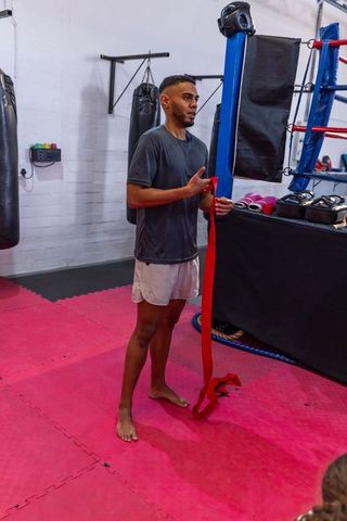 Young asian boxer wrapping red handwraps while standing by ring on pink training mats
