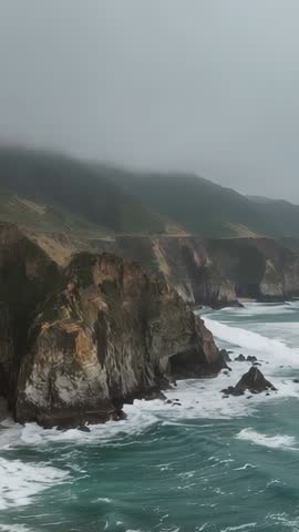 Vertical drone filming rugged coastal cliffs and crashing surf under misty overcast sky