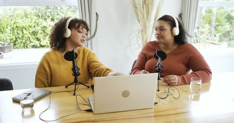 Diverse female podcast hosts recording together at home wearing headphones and sweaters