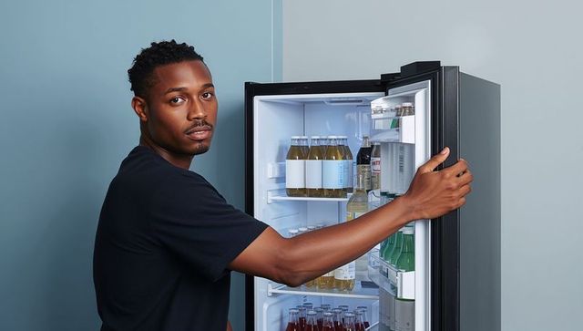 Man holding fridge door open with illuminated beverage bottles, minimalist studio backdrop