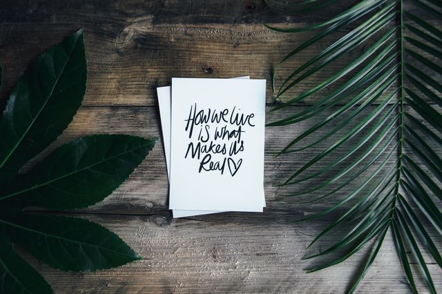 Handwritten quote on card placed on rustic wooden surface surrounded by green leaves. Perfect for wellness blogs, motivational social media posts, and lifestyle websites looking to inspire their audience. Ideal for use in promotional materials for stationary products or nature-theme designs.