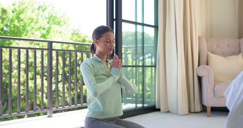 African american woman meditating at home by sunlit balcony and sliding glass doors