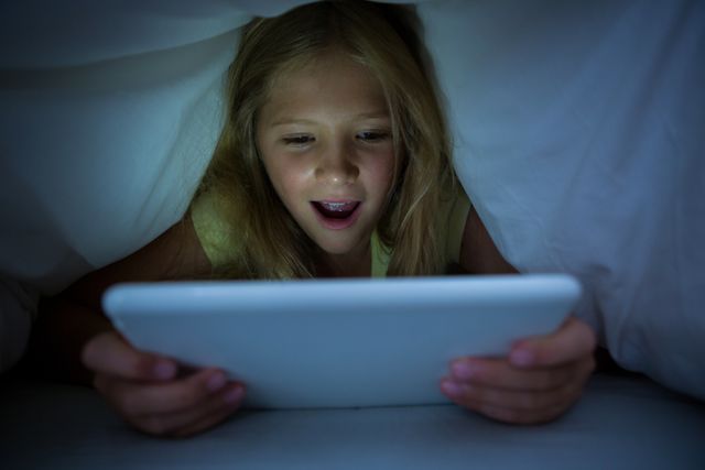 Young girl lying in bed under a blanket, using a tablet with a happy expression. Ideal for illustrating concepts of childhood, technology use, bedtime routines, and digital entertainment. Suitable for articles, blogs, and advertisements related to children's technology use, parenting, and nighttime activities.