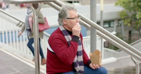 Older man eating takeout on urban overpass stairs during lunch break, commuters passing