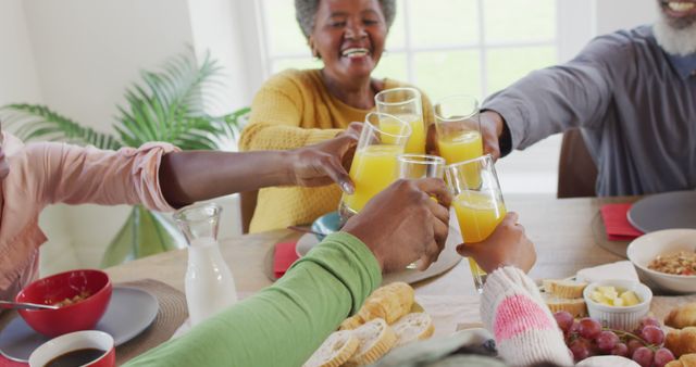 Multi-generational family toasting with glasses of orange juice around breakfast table. Ideal for usage in family-centric advertisements, healthy living promotions, and community-related content. Perfect for illustrating the joy of togetherness and healthy eating. Depicts happiness and bonding in domestic setting.