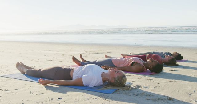 Group of women lying on mats on a sandy beach performing yoga during sunrise, promoting relaxation and wellness. Useful for websites related to fitness, wellness retreats, outdoor activities, meditation guides, and healthy lifestyle inspiration.