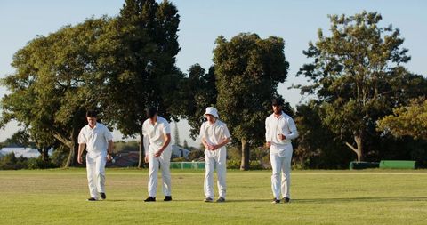 Four cricket players walking on sunlit suburban field wearing white kit and caps
