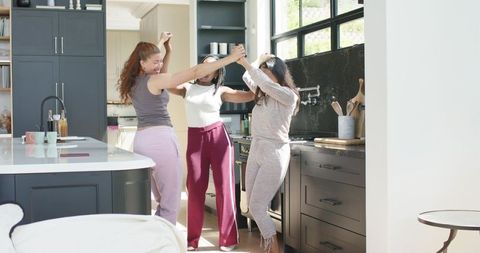 African american women dancing and laughing in sunlit kitchen holding hands, casual loungewear
