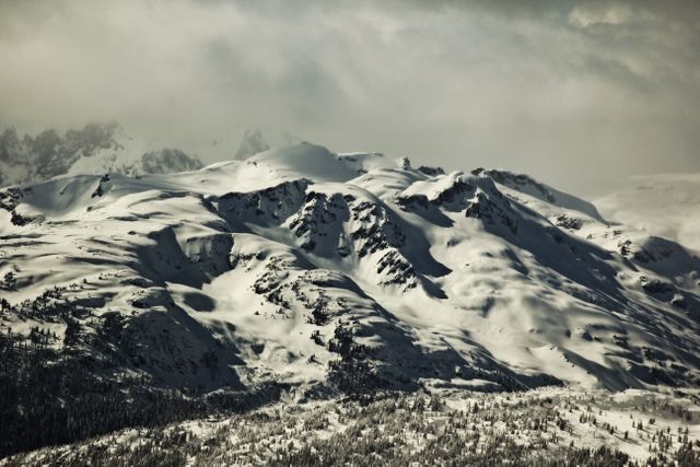 Snow-covered mountain range under a cloudy sky, with rugged peaks and valleys. Ideal for use in articles or projects related to winter landscapes, adventure, nature exploration, travel destinations, climate, and serene natural beauty.