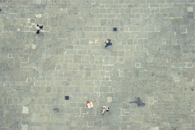 Aerial view shows several people walking on a stone-paved square with a grid pattern. Individuals appear small and scattered across the scene, creating a sense of scale and space. Useful for urban life, cityscapes, and architecture themes. Suitable for websites, advertisements, travel blogs, or city planning presentations.