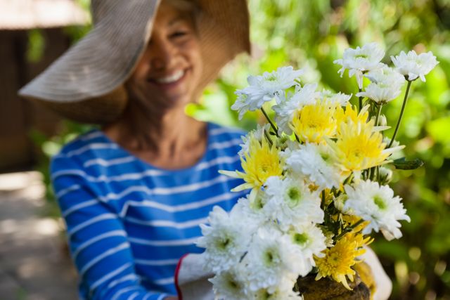Smiling senior woman wearing hat holding fresh flowers at backyard