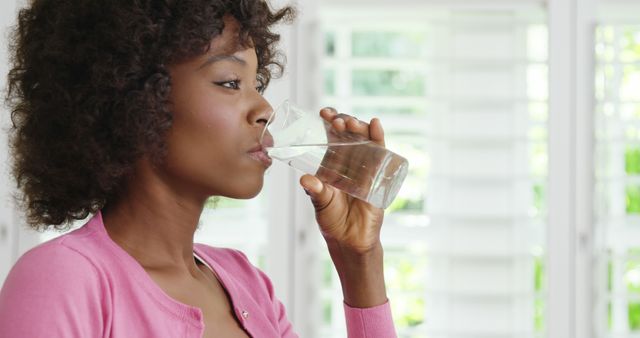 Woman in pink sweater enjoying a glass of water while standing indoors, with natural light streaming through windows. Useful for themes of hydration, self-care, healthy living, and daily routines, especially for articles or content related to wellness, health tips, and lifestyle.