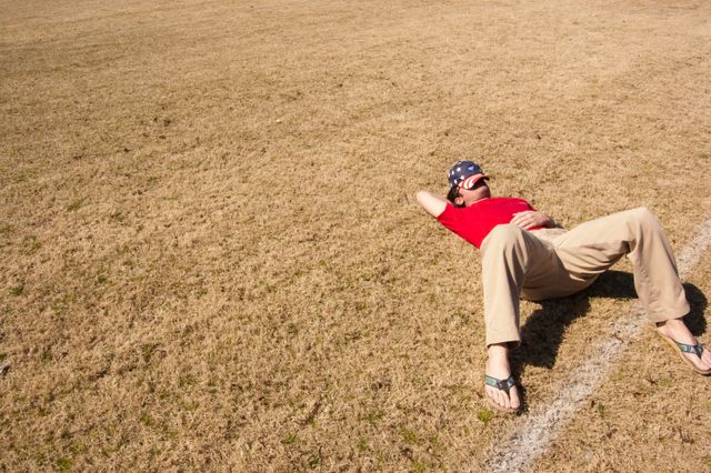 This image shows a man relaxing on a dry grass field under bright sunlight. He appears to be enjoying a peaceful moment outdoors, dressed in casual clothing including a red shirt, beige pants, and flip-flops. Ideal for illustrating leisure, outdoor relaxation, summer activities, or casual lifestyle contexts.