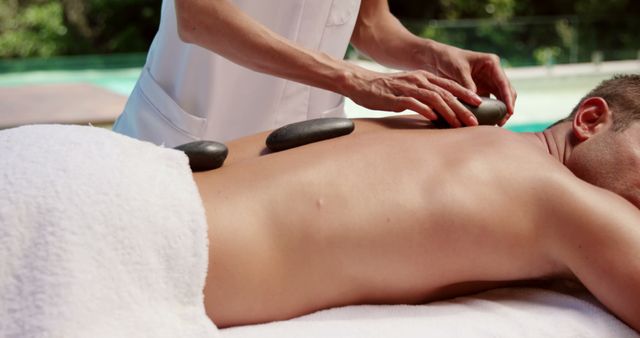 Scene depicts a man receiving a hot stone massage at an outdoor spa. Useful for promoting spa services, relaxation therapies, and wellness retreats. Can be used in advertisements and websites related to health and wellness products and services.