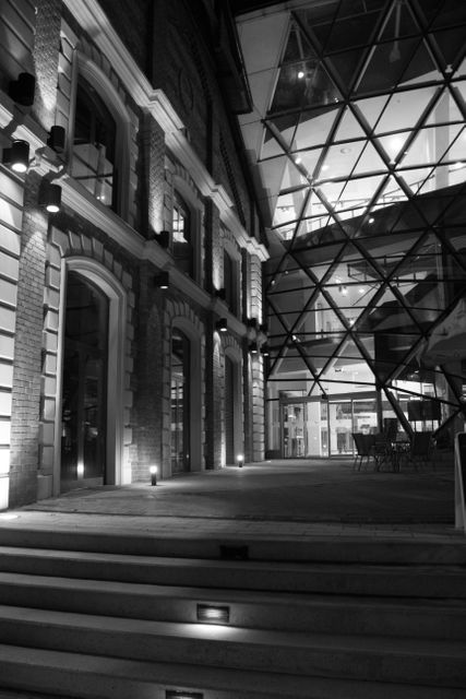 This image captures a dramatic night view of a modern glass structure adjoining a historic brick building. The stark contrast between the sleek glass facade and the classic brickwork is accentuated by soft lighting. This versatile contrast makes the image ideal for architectural presentations, urban development projects, historical versus contemporary comparisons, and promotional materials for hotels or commercial real estate showcasing unique design elements.