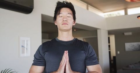 Asian man meditating at home under skylight doing namaste pose for mindfulness wellness