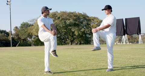 Two cricketers stretching on grassy outfield in white kit and caps during daylight training