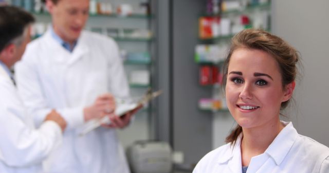 A young Caucasian female pharmacist smiles at the camera, with two male colleagues discussing in the background, with copy space. Her professional demeanor in a pharmacy setting suggests a collaborative work environment focused on healthcare.