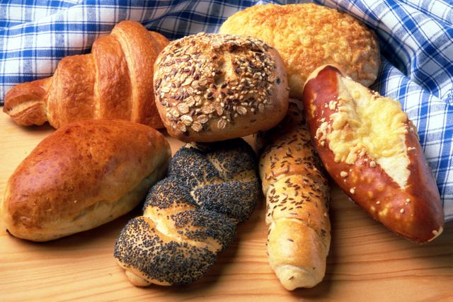 Assorted freshly baked breads and pastries including croissant, pretzel, and seeded rolls displayed on a wooden table with a blue and white checkered cloth. Perfect for illustrating articles or advertisements for bakeries, breakfast menus, and food blogs.