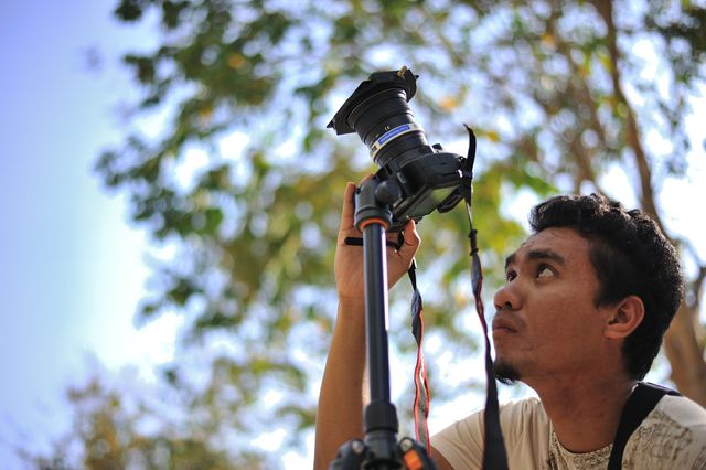 Man adjusting camera on tripod during outdoor shoot under trees, ideal for illustrating photography practice, nature-focused photography, or showcasing photography equipment.