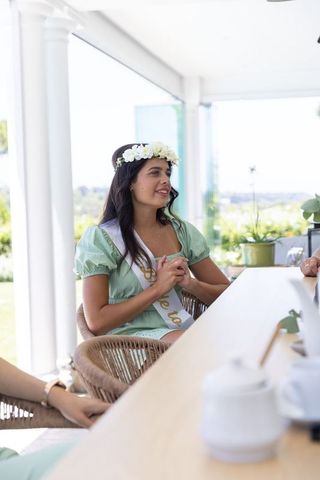 Bride-to-be sitting on sunlit veranda wearing floral crown and white sash at bridal shower