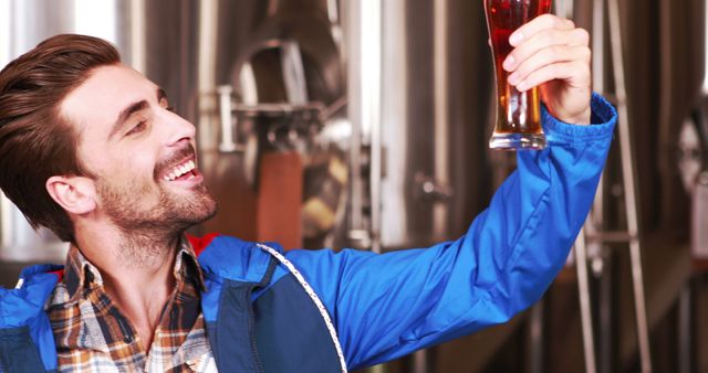 Man with a plaid shirt and blue jacket inspects beer quality in a brewery. Industrial equipment is visible in the background. Suitable for use in articles or advertisements related to breweries, craft beer, beer production processes, or quality control measures in beverage manufacturing.