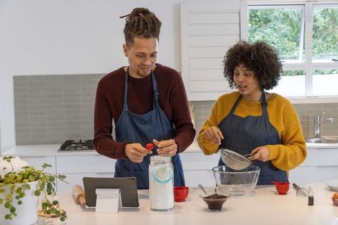 Diverse couple baking in modern kitchen measuring flour, sifting, checking tablet recipe