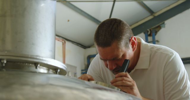 Brewmaster is closely inspecting a large stainless steel fermentation tank in a brewery. He is looking attentively with a flashlight to ensure the quality and accuracy of the brewing process. Perfect for use in articles, advertisements, and marketing materials related to craft beer production, quality control, and industrial processes in microbreweries.