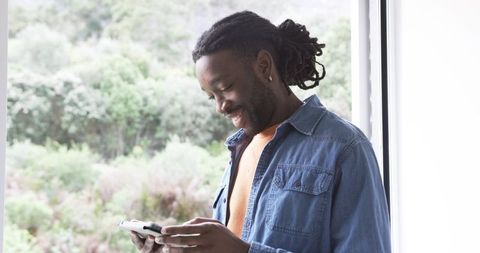 African American man standing by window smiling while using smartphone in bright interior