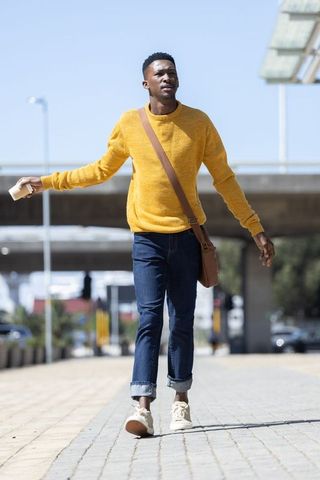 African man walking with coffee wearing mustard sweater carrying brown messenger bag