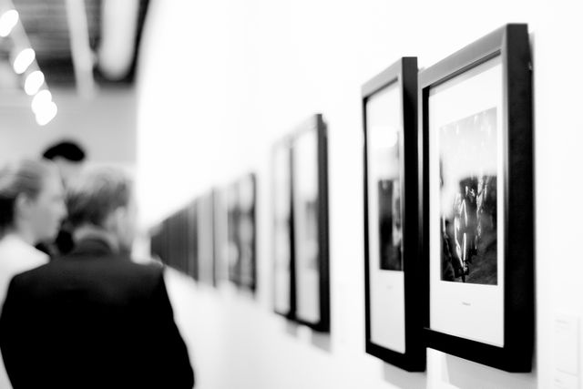 Image depicts visitors viewing framed photographs on the wall in a modern art gallery. This can be used to depict art exhibitions, cultural events, creative atmospheres, or for promoting art shows. It showcases a high-contrast black and white aesthetic that adds a timeless feel to the scene.