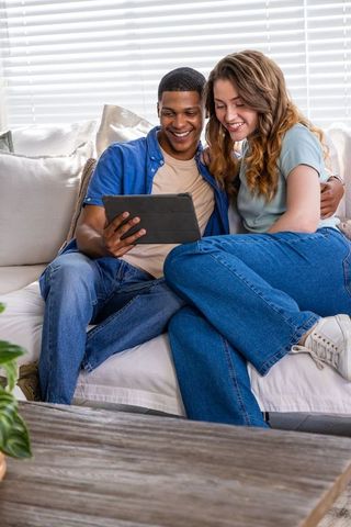 Young interracial couple sharing tablet on sofa, cozy denim outfits, smiling together