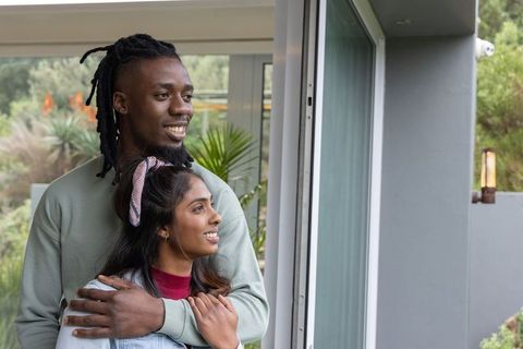 African American man and Indian woman embracing by sliding glass door with garden view