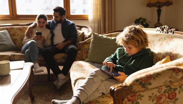 Child using tablet on vintage floral sofa in cozy sunlit living room with parents