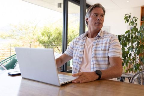 Mature man working remotely on laptop, sitting at table with balcony view and plants