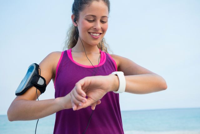Young woman enjoying a day at the beach while using her smart watch. She is smiling and appears to be in a good mood, possibly tracking her fitness progress or checking notifications. Ideal for use in advertisements for fitness technology, healthy lifestyle promotions, or summer activities.