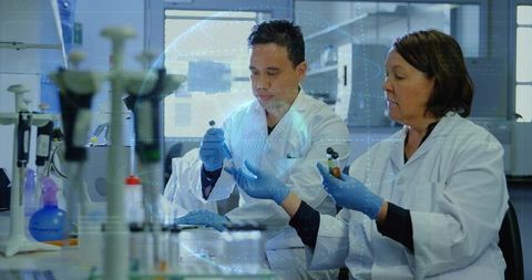 Senior scientist guiding trainee holding sample vials and using pipettes in modern lab