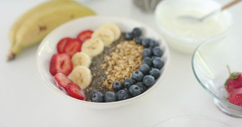 Serving breakfast bowl with granola, chia, banana, strawberries and blueberries on white table