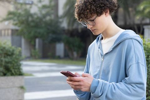 Teen texting on campus pathway wearing blue hoodie and glasses, modern student technology