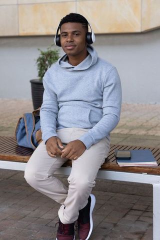 Urban teen student wearing headphones and hoodie sitting on bench with backpack, notebook