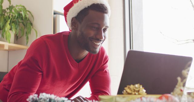 Man wearing Santa hat engaging in a virtual celebration using laptop. Bright, festive atmosphere with Christmas decorations. Ideal for holiday marketing campaigns, festive website content, virtual holiday event promotions, and technological advancements in virtual gatherings. Showcases use of technology in holiday celebrations.