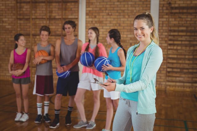 Smiling coach holding clipboard in basketball court with team of young players holding basketballs. Ideal for use in sports training programs, youth coaching advertisements, fitness and health campaigns, and educational materials promoting teamwork and leadership.