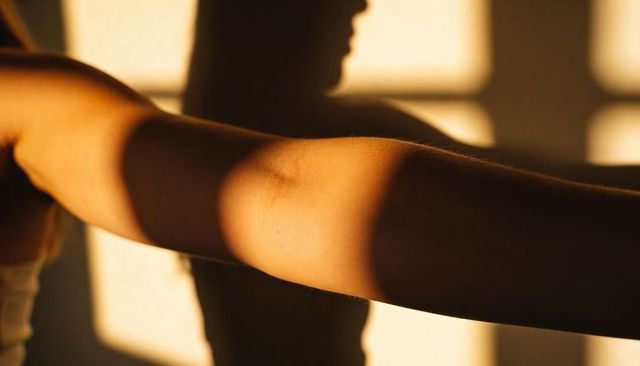 Sunlit forearm extending across shadowed torso with window grid and golden warmth