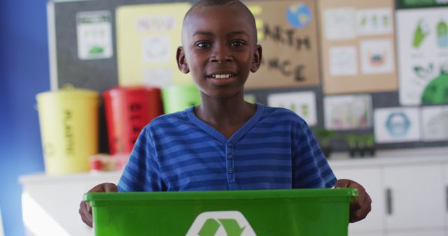 African american schoolboy smiling, holding recycling bin, standing in classroom. children at primary school in summer.